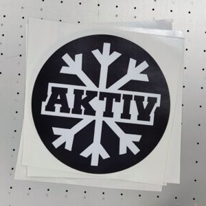 Rundt sort klistremerke med hvit snøkrystall og teksten "AKTIV" i midten, 15 cm diameter.