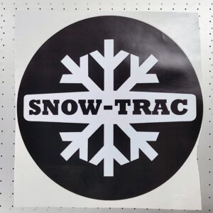Rundt Snow-Trac klistremerke med hvit snøkrystall og tekst på sort bakgrunn, 30 cm diameter, vist på hvit monteringsflate.